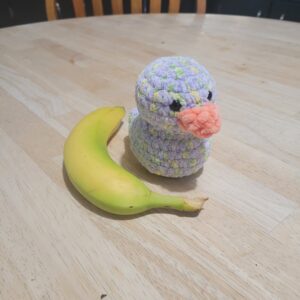 A lavender crochet duck sitting on a wood table beside a banana.